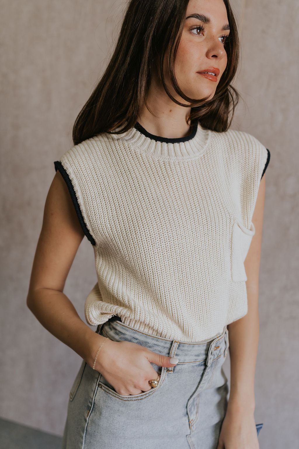Side view of female model wearing the Vivian Ivory & Navy Knit Sleeveless Sweater which features Cream Cable Knit Fabric, Navy Trim Details, Left Front Chest Pocket, High Neckline and Sleeveless.