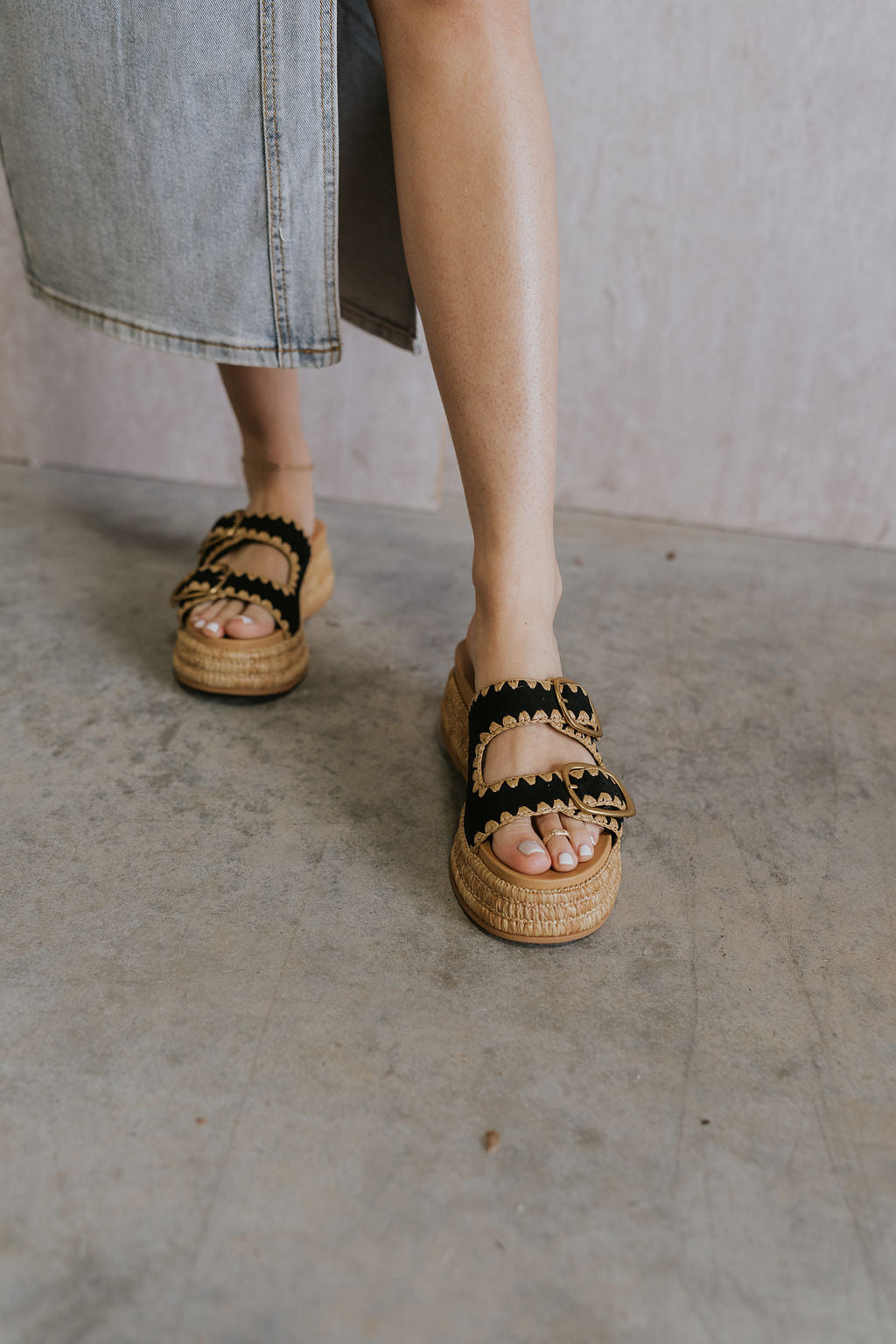 Front view of female model wearing the Wanika Sandal in Onyx which features black onyx nubuck fabric, raffia platform sole, raffia pattern details, rubber outsole, adjustable gold buckle two straps and round toe