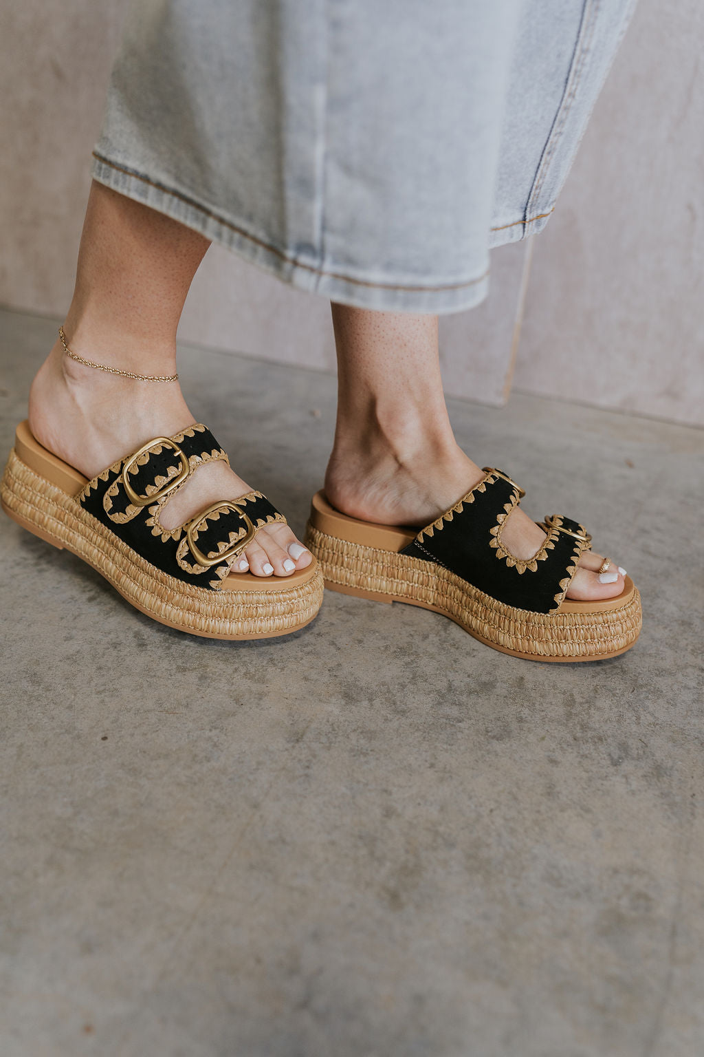 Close up view of female model wearing the Wanika Sandal in Onyx which features black onyx nubuck fabric, raffia platform sole, raffia pattern details, rubber outsole, adjustable gold buckle two straps and round toe