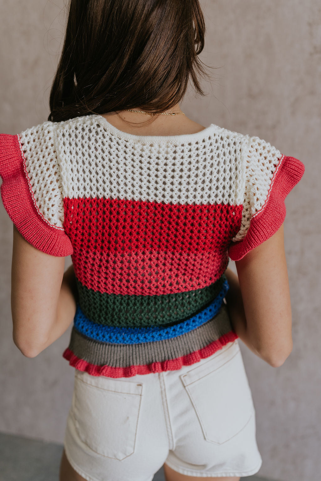 Back view of female model wearing the Andrea Pink Multi Knit Ruffle Top which features Blue, Green, Pink, Grey and Cream Open Knit Fabric, Ruffle Hem, V-Neckline and Short Sleeves