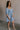 Full body side view of female model wearing the Hallie Light Chambray Mini Dress which features Light Chambray Tencel Fabric, Mini Length, Square Neckline, Half Sleeves with Ruffle Hem Details and Smocked Back