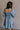 Back view of female model wearing the Hallie Light Chambray Mini Dress which features Light Chambray Tencel Fabric, Mini Length, Square Neckline, Half Sleeves with Ruffle Hem Details and Smocked Back