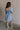Full body back view of female model wearing the Hallie Light Chambray Mini Dress which features Light Chambray Tencel Fabric, Mini Length, Square Neckline, Half Sleeves with Ruffle Hem Details and Smocked Back
