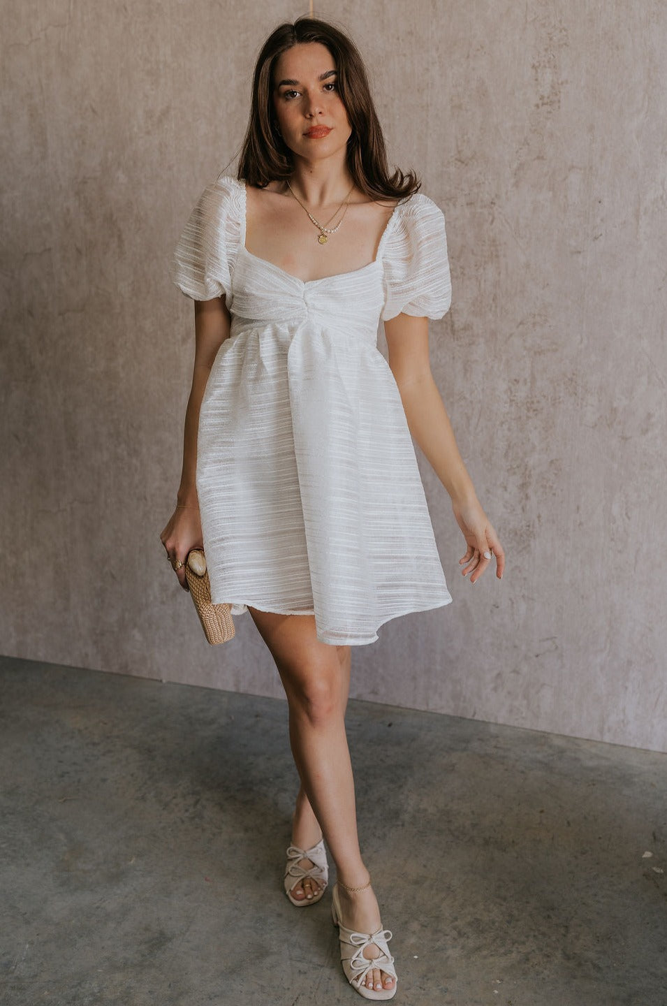 Full body view of female model wearing the Julia Off White Short Puff Sleeve Mini Dress which features Off White Stripe Sheer Fabric, White Lining, Mini Length, Ruched Upper, Sweetheart Neckline, Short Puff Sleeves and Back Cut-Out Detail with Zipper and Tie Closure