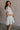 Side view of female model wearing the Julia Off White Short Puff Sleeve Mini Dress which features Off White Stripe Sheer Fabric, White Lining, Mini Length, Ruched Upper, Sweetheart Neckline, Short Puff Sleeves and Back Cut-Out Detail with Zipper and Tie Closure
