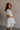 Side view of female model wearing the Julia Off White Short Puff Sleeve Mini Dress which features Off White Stripe Sheer Fabric, White Lining, Mini Length, Ruched Upper, Sweetheart Neckline, Short Puff Sleeves and Back Cut-Out Detail with Zipper and Tie Closure