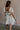 Back view of female model wearing the Julia Off White Short Puff Sleeve Mini Dress which features Off White Stripe Sheer Fabric, White Lining, Mini Length, Ruched Upper, Sweetheart Neckline, Short Puff Sleeves and Back Cut-Out Detail with Zipper and Tie Closure