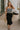 Full body front view of model wearing the Arabella Black Wrap Midi Skirt that has black lightweight fabric, a wrap front with a side tie detail, and back slit. Worn with striped tube top.