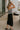 Full body side view of model wearing the Arabella Black Wrap Midi Skirt that has black lightweight fabric, a wrap front with a side tie detail, and back slit. Worn with striped tube top.