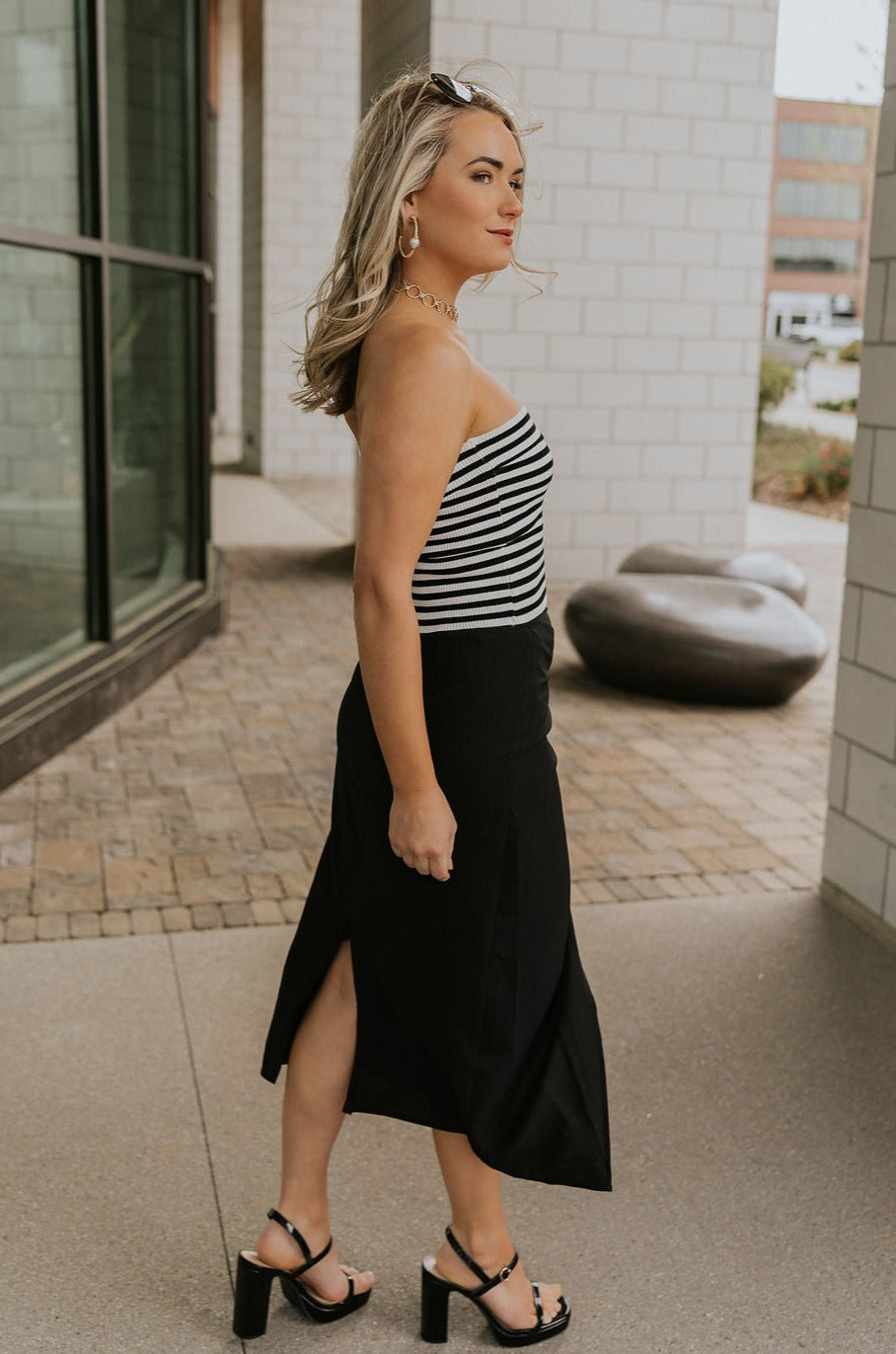 Full body side view of model wearing the Arabella Black Wrap Midi Skirt that has black lightweight fabric, a wrap front with a side tie detail, and back slit. Worn with striped tube top.