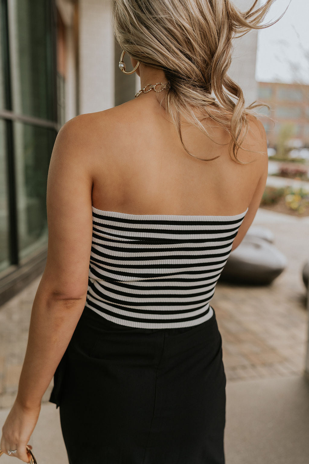 Upper body back view of female model wearing the Londyn White & Black Striped Tube Top that has horizontal white and black stripes and a straight strapless neckline. Worn with black skirt.