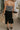 Back view of model wearing the Arabella Black Wrap Midi Skirt that has black lightweight fabric, a wrap front with a side tie detail, and back slit. Worn with striped tube top.