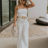 Full body front view of blonde model wearing the Regan Wide Leg Denim Jumpsuit in White, that has white denim fabric, wide legs, and thin straps. Styled with a beige belt. Model is holding beige purse.
