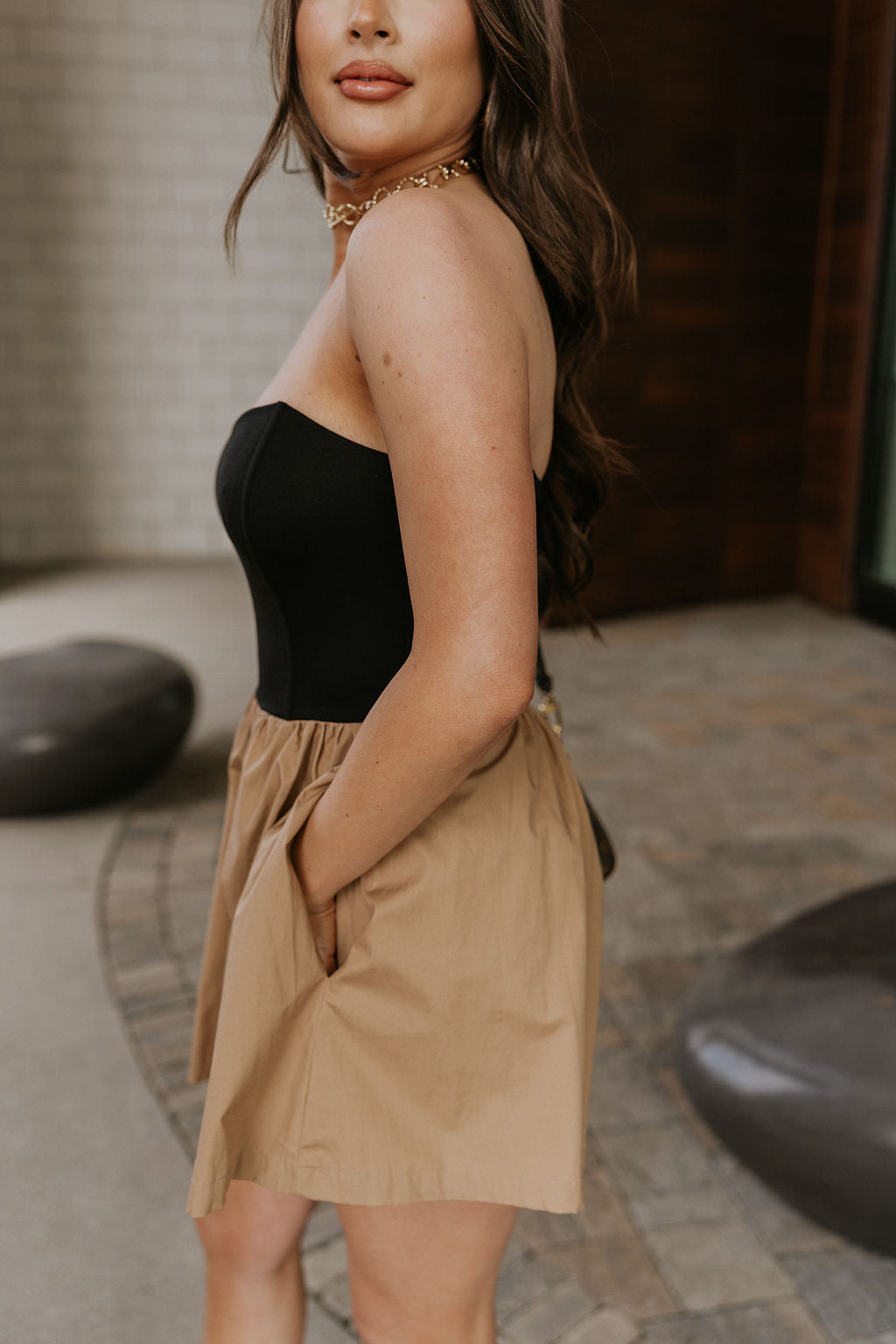 Side view of female model wearing the Parker Black & Mocha Strapless Romper that has a black upper with a strapless neck attached to mocha shorts with a wide fit and pockets.