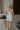 Full body back view of female model wearing the Emerson White Blue Floral Ruffle Mini Dress which features White Lightweight Fabric, Blue Floral Print, Ruffle Hem Design, Square Neckline and sleeveless