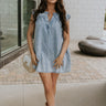 Full body front view of female model wearing the Arya Denim Mini Dress that has light wash denim, sleeveless body, mini length, and a notched neck with a tie closure. 