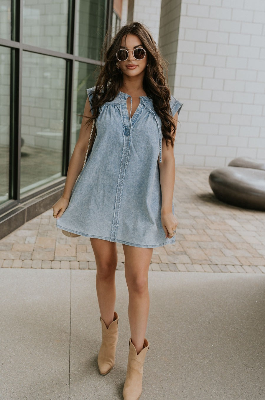 Full body front view of female model wearing the Arya Denim Mini Dress that has light wash denim, sleeveless body, mini length, and a notched neck with a tie closure. 