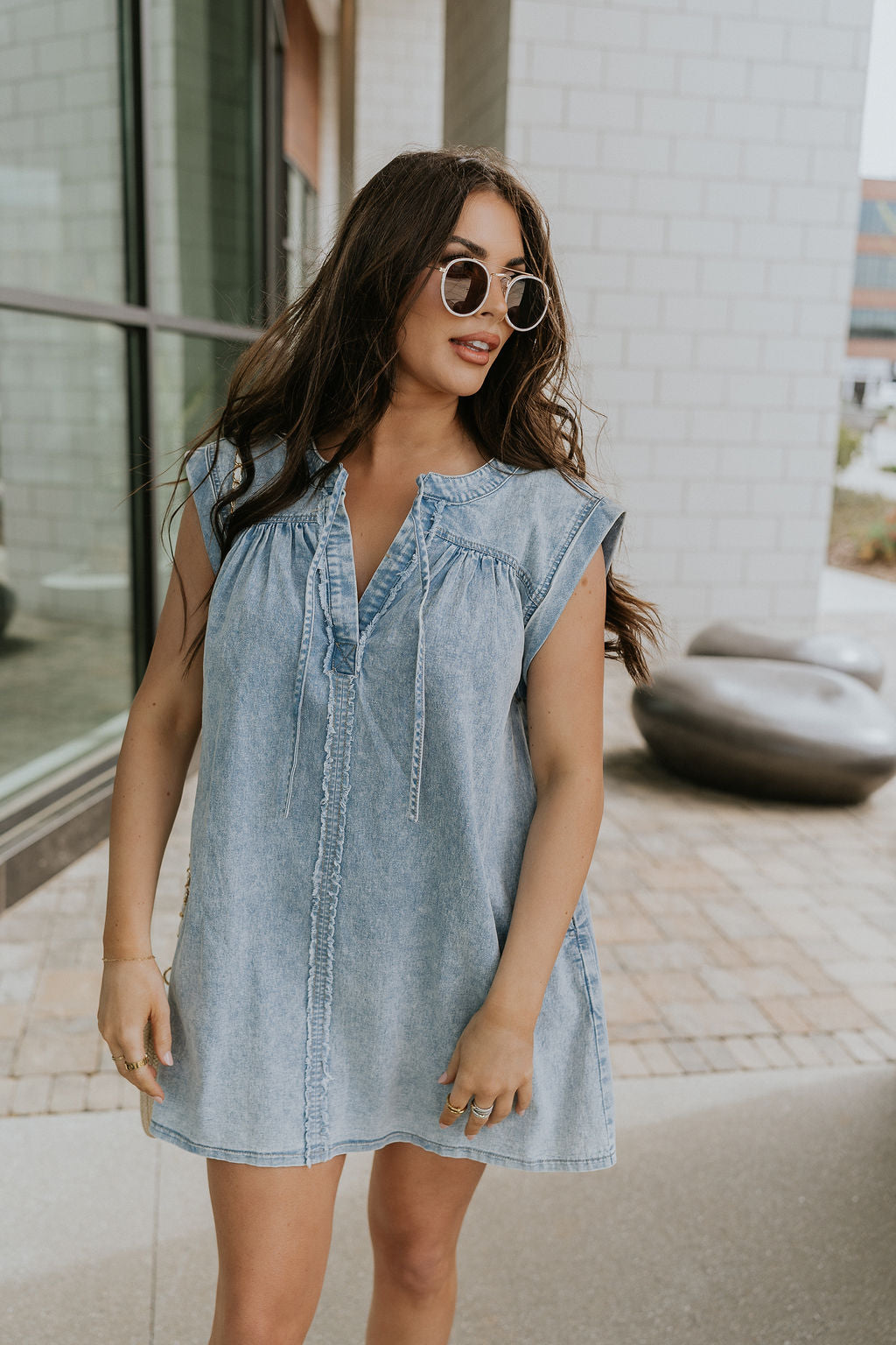 Front view of female model wearing the Arya Denim Mini Dress that has light wash denim, sleeveless body, mini length, and a notched neck with a tie closure. 