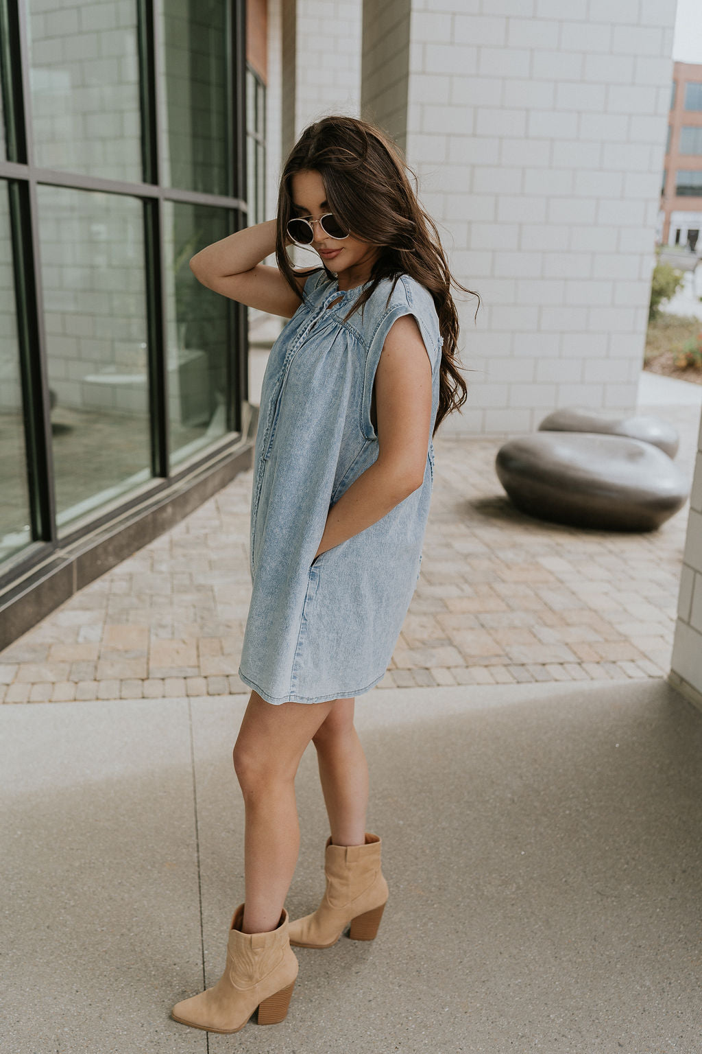 Full body side view of female model wearing the Arya Denim Mini Dress that has light wash denim, sleeveless body, mini length, and a notched neck with a tie closure. 
