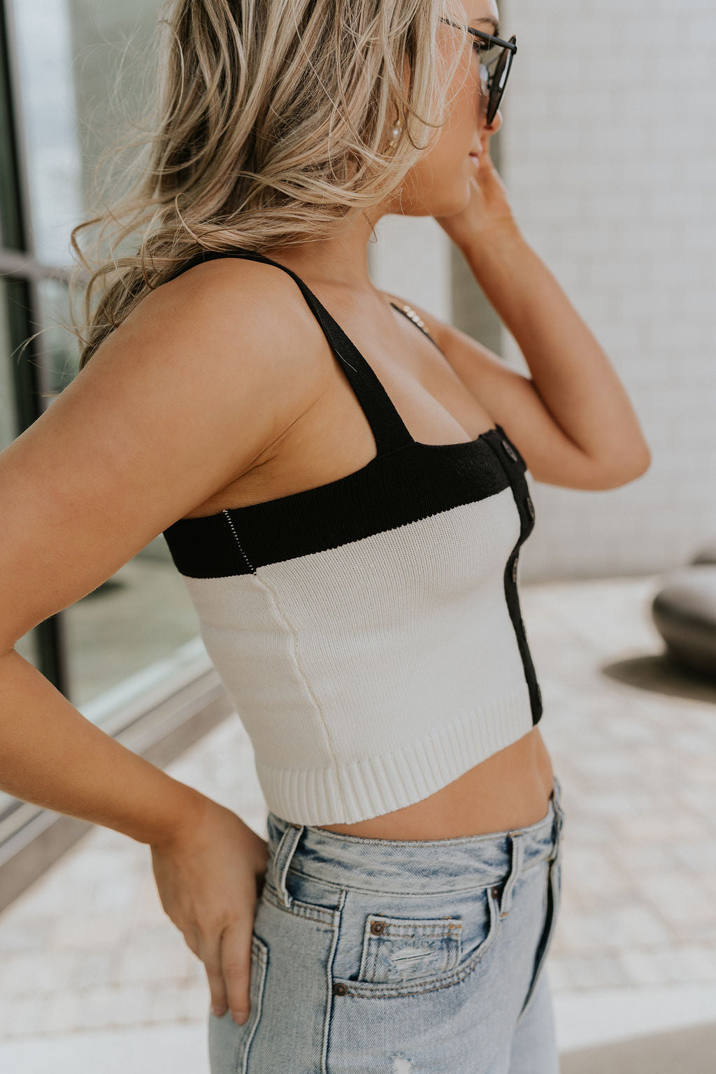 Upper body side view of female model wearing the Amara Cream & Black Cropped Tank that has knit fabric with cream and black colorblocking and faux black buttons. Worn with blue jeans.