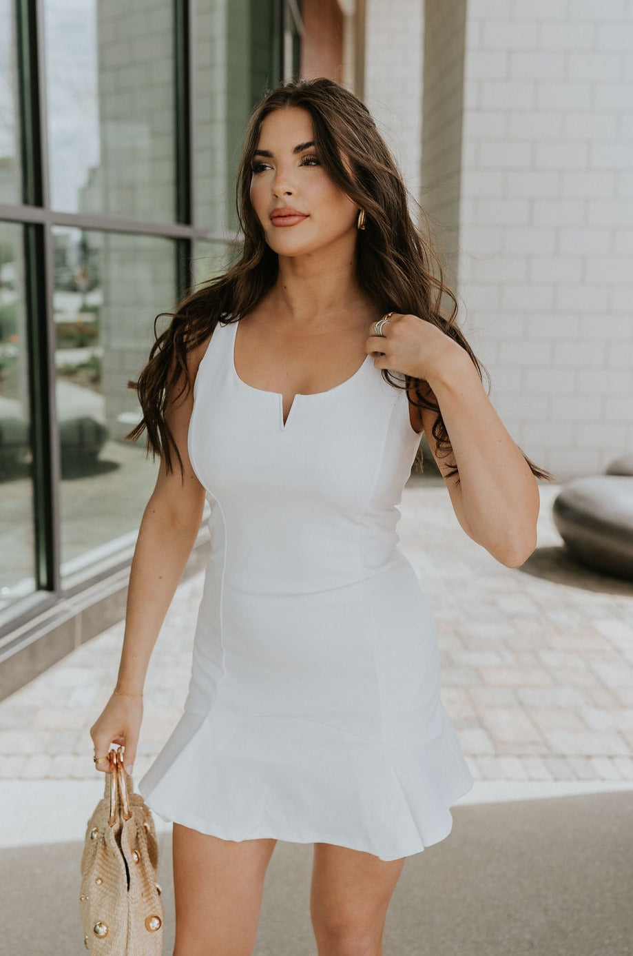 Front view of female model wearing the Isabel White Sleeveless Mini Dress that has white fabric, a notched neckline, mini length, and sleeveless bodice. Model is holding beige purse.