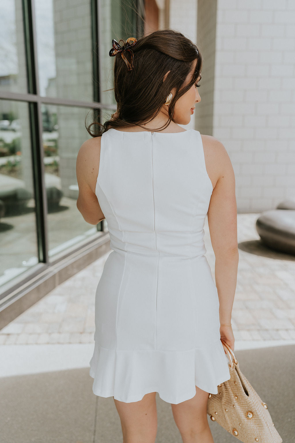 Back view of female model wearing the Isabel White Sleeveless Mini Dress that has white fabric, a notched neckline, mini length, and sleeveless bodice. Model is holding beige purse.