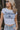 Close front view of female model wearing the Brooklyn White Short Sleeve Graphic Tee that has a navy and light blue graphic that says "EST. 1968 Brooklyn, Greatest Performance Central Division, New York, Western Academy, Campus Sports League" with a star and angel wings. Worn with blue jeans.