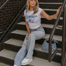 Female model is shown sitting on stairs while wearing the Brooklyn White Short Sleeve Graphic Tee that has a navy and light blue graphic that says "EST. 1968 Brooklyn, Greatest Performance Central Division, New York, Western Academy, Campus Sports League" with a star and angel wings. Worn with blue jeans.