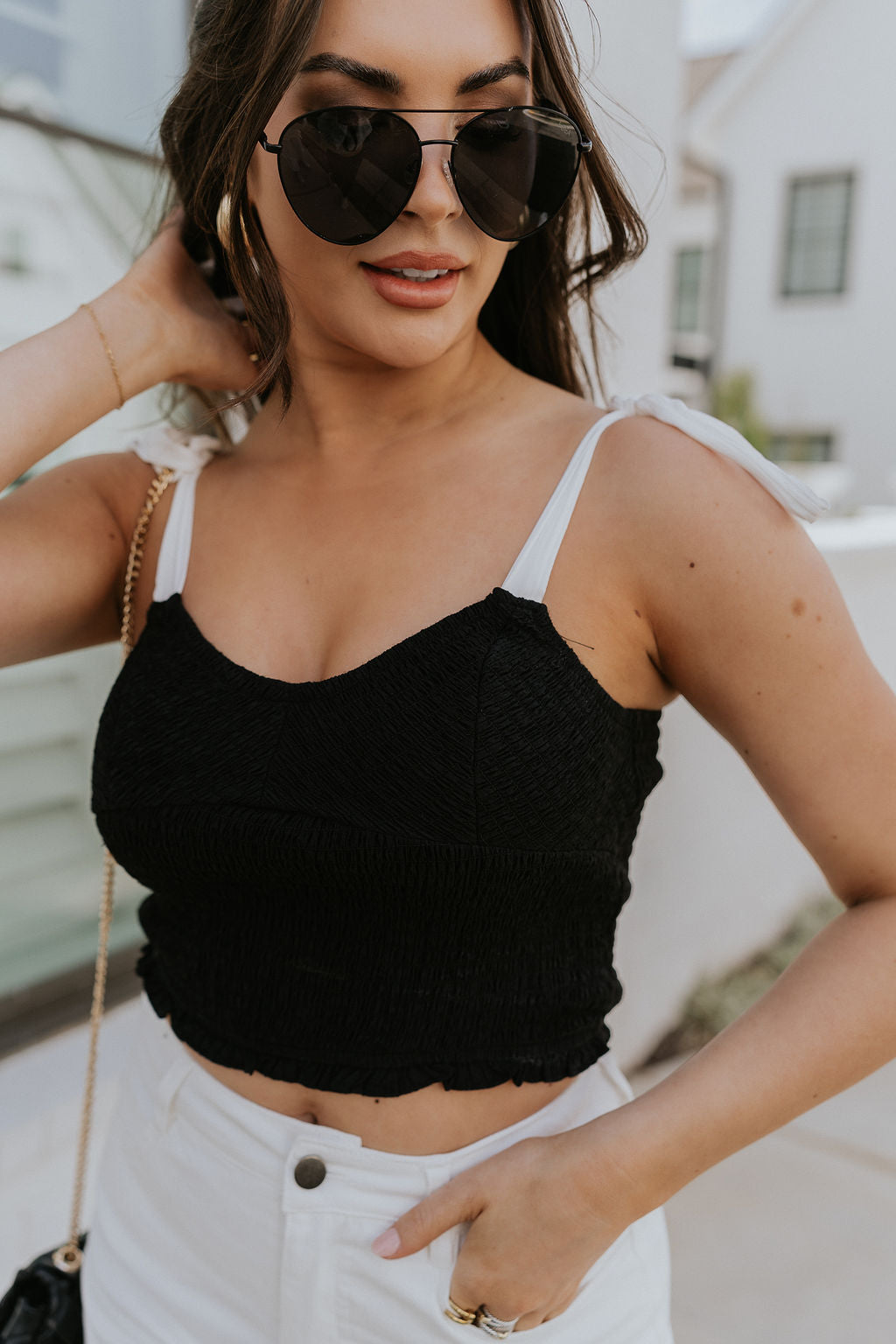 Close front view of female model wearing the Reese Black & White Cropped Tank Top that has black smocked body, white tie straps and a cropped waist. Worn with white pants.