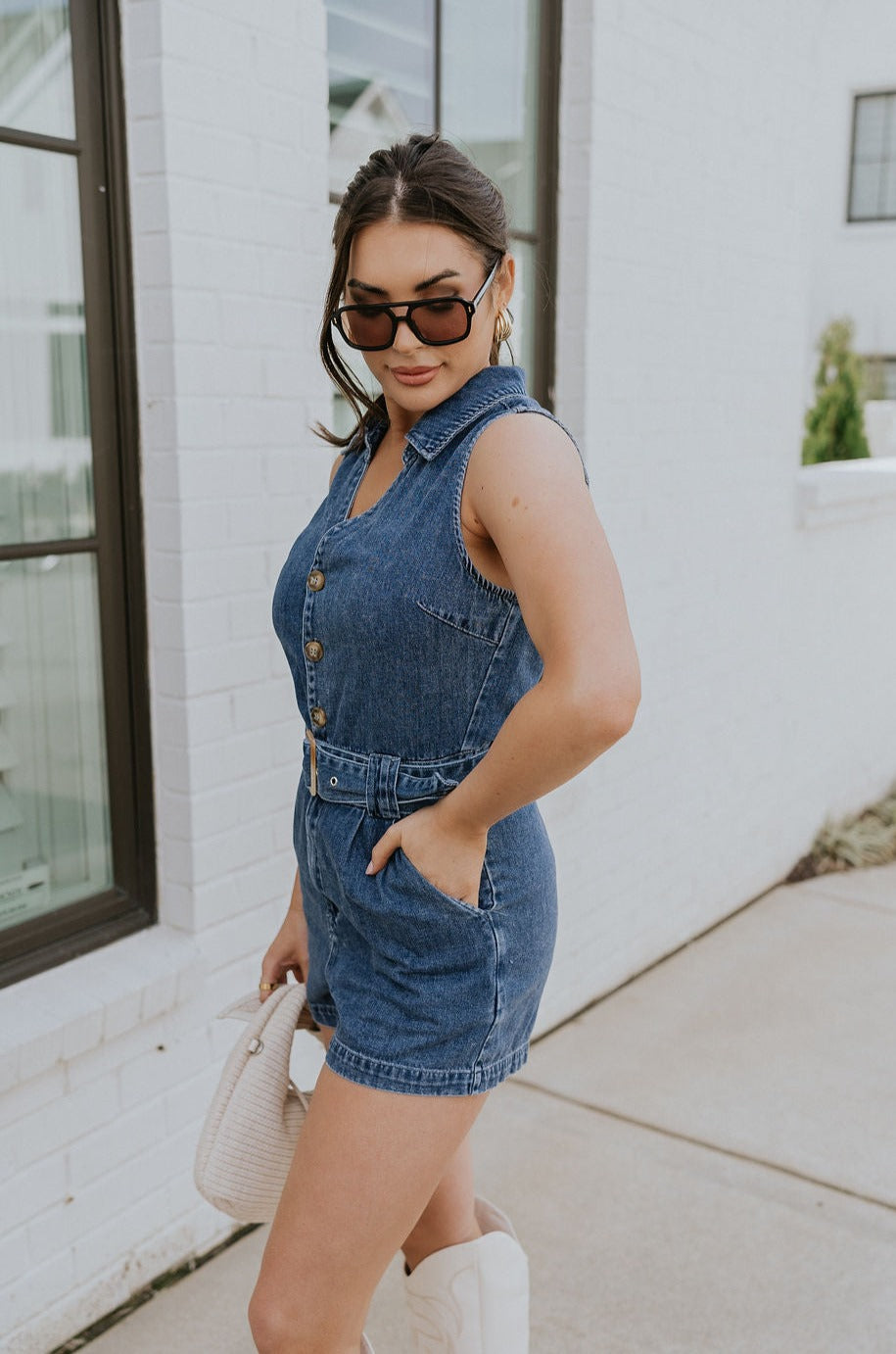 Side view of female model wearing the Georgia Belted Denim Romper in medium wash, that has a button up front, collar, and belted waist. Paired with white boots.