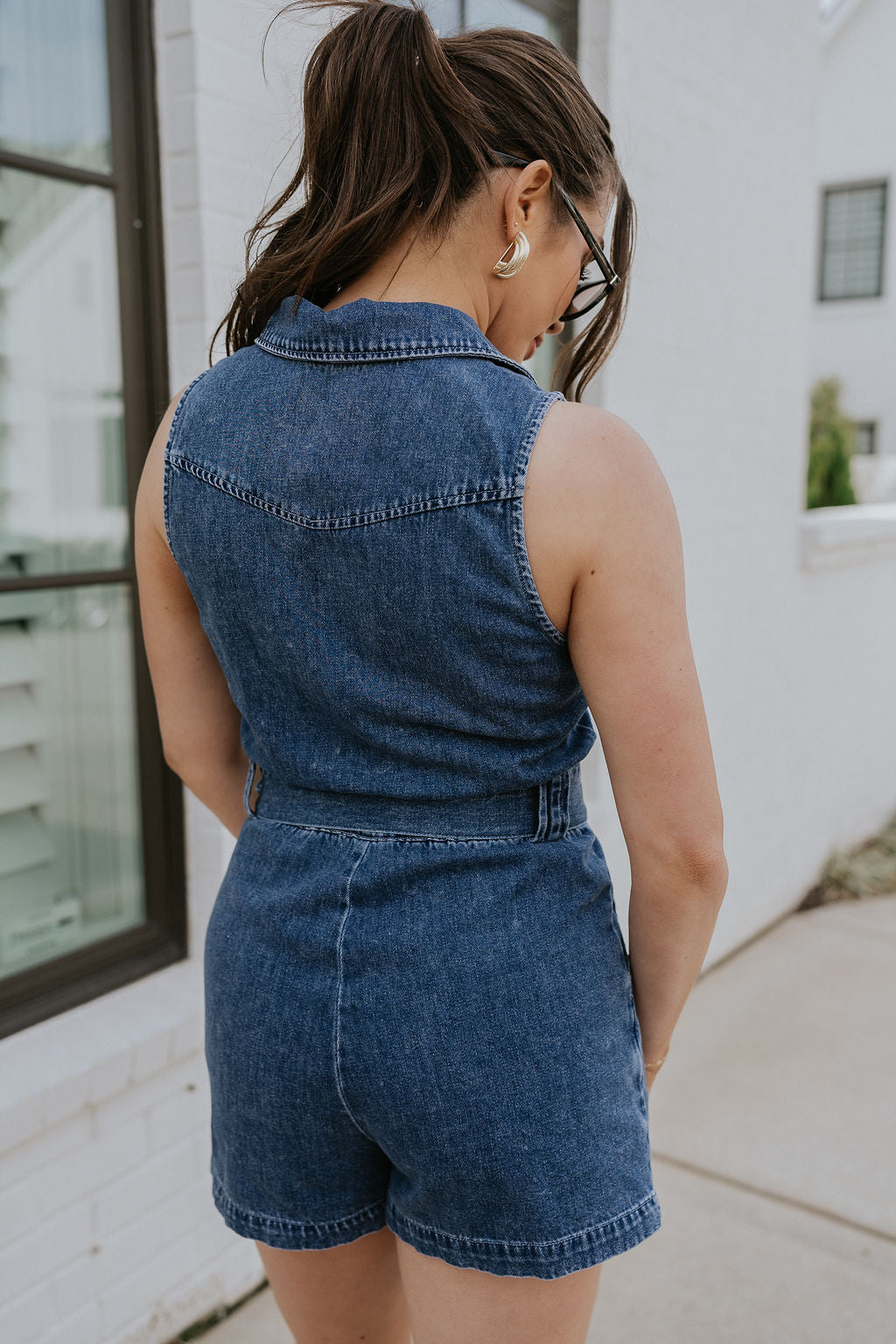 Back view of female model wearing the Georgia Belted Denim Romper in medium wash, that has a button up front, collar, and belted waist. 