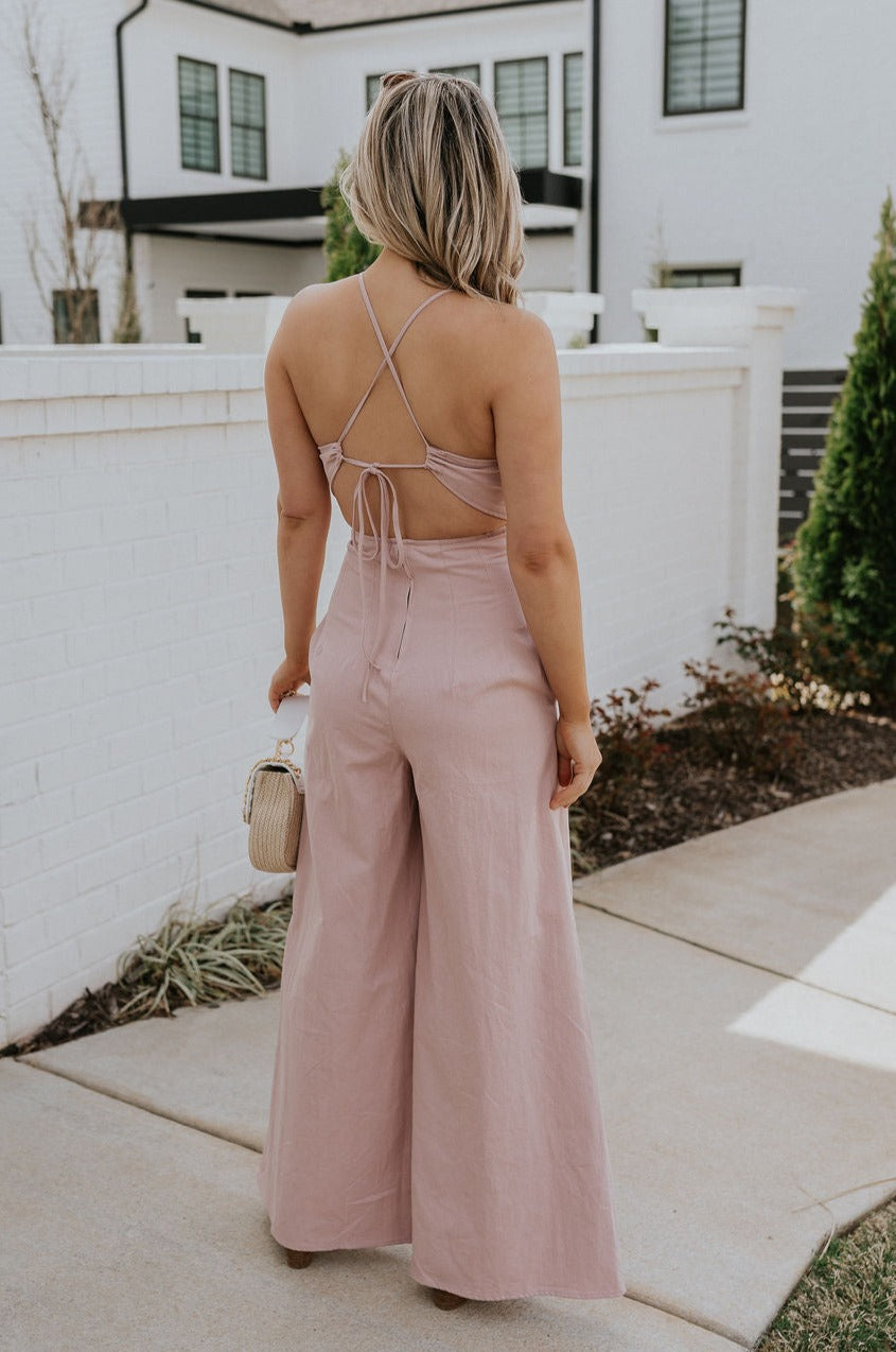 Full body back view of female model wearing the Aleah Mauve Pink Sleeveless Jumpsuit which features Mauve Pink Lightweight Fabric, Wide Leg Pants, Upper Ruched Detail, Two Side Pockets, Scooped Neckline, Adjustable Straps and Open Back with Tie Closure and Zipper