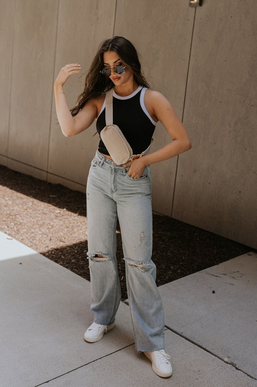 Full body front view of female model wearing the Ari Black & White Brami that has black ribbed fabric with white trim and a round neck. Paired with blue jeans and beige sling bag.