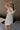 Side view of female model wearing the Rhea White Floral Ruffle Mini Dress which features Off White Lightweight Fabric, Mini Babydoll Style, Off White Lining, Monochrome Floral Print with Silver Metallic Thread, Sweetheart Neckline, Ruffle Straps and Open Back with Tie Closure