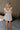 Full body back view of female model wearing the Rhea White Floral Ruffle Mini Dress which features Off White Lightweight Fabric, Mini Babydoll Style, Off White Lining, Monochrome Floral Print with Silver Metallic Thread, Sweetheart Neckline, Ruffle Straps and Open Back with Tie Closure