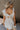 Back view of female model wearing the Rhea White Floral Ruffle Mini Dress which features Off White Lightweight Fabric, Mini Babydoll Style, Off White Lining, Monochrome Floral Print with Silver Metallic Thread, Sweetheart Neckline, Ruffle Straps and Open Back with Tie Closure
