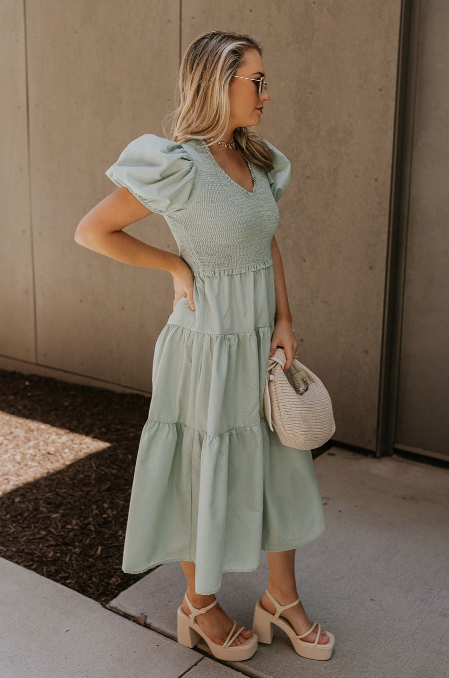 Full body side view of female model wearing the Sloan Sage Puff Sleeve Midi Dress which features Light Sage Lightweight Fabric, Light Sage Lining, Midi Length, Tiered Skirt, Smocked Upper, V-Neckline and Short Puff Sleeves