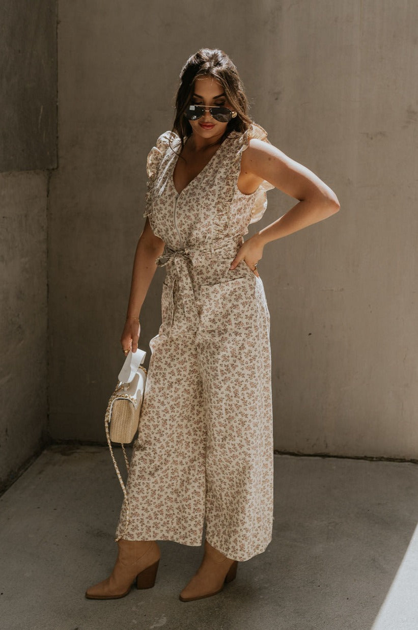 Full body view of female model wearing the Myla Cream Pink Floral Sleeveless Jumpsuit which features Cream Cotton Fabric, Pink Floral Print, Wide Leg Pants, Tie Strap with Belt Loops, V-Neckline and Ruffle Straps