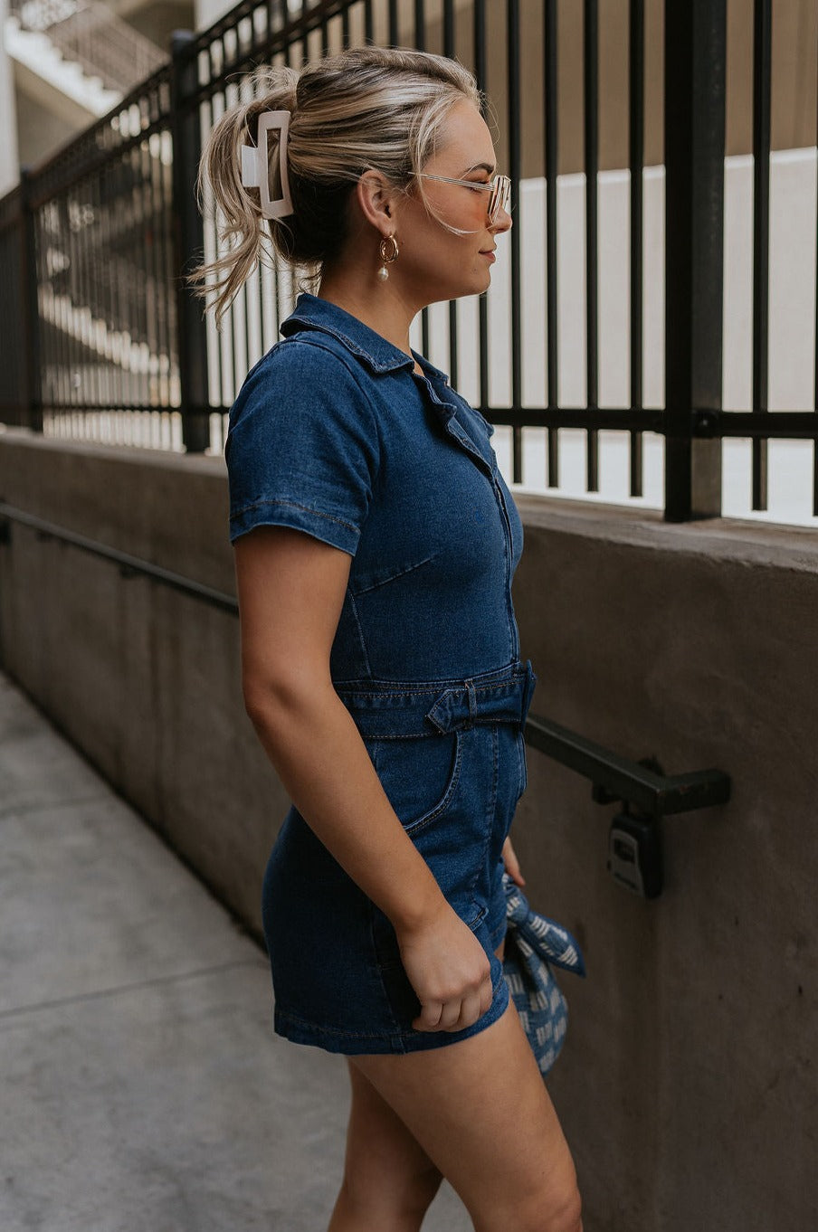 Side view of female model wearing the Mavis Denim Short Sleeve Zip-Up Romper which features Denim Fabric, Two Front Pockets, Front Zipper Closure, Adjustable Monochrome Belt with Loops, Collared Neckline and Short Sleeves
