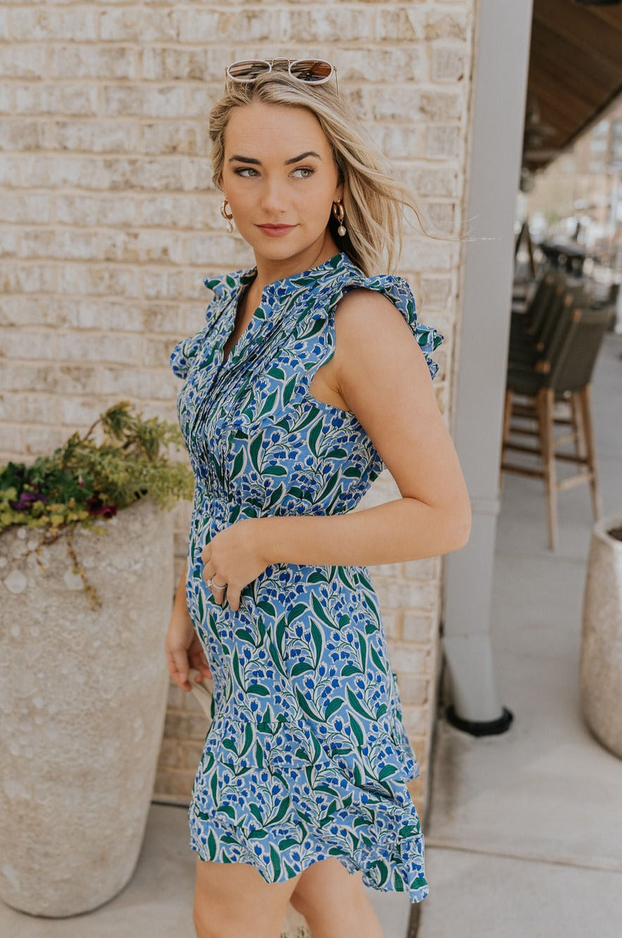 Side view of female model wearing the June Blue & Green Floral Ruffle Mini Dress which features Blue and Green Floral Print, Ruffle Tiered Design, Mini Length, High Neck with V-Cutout and Sleeveless