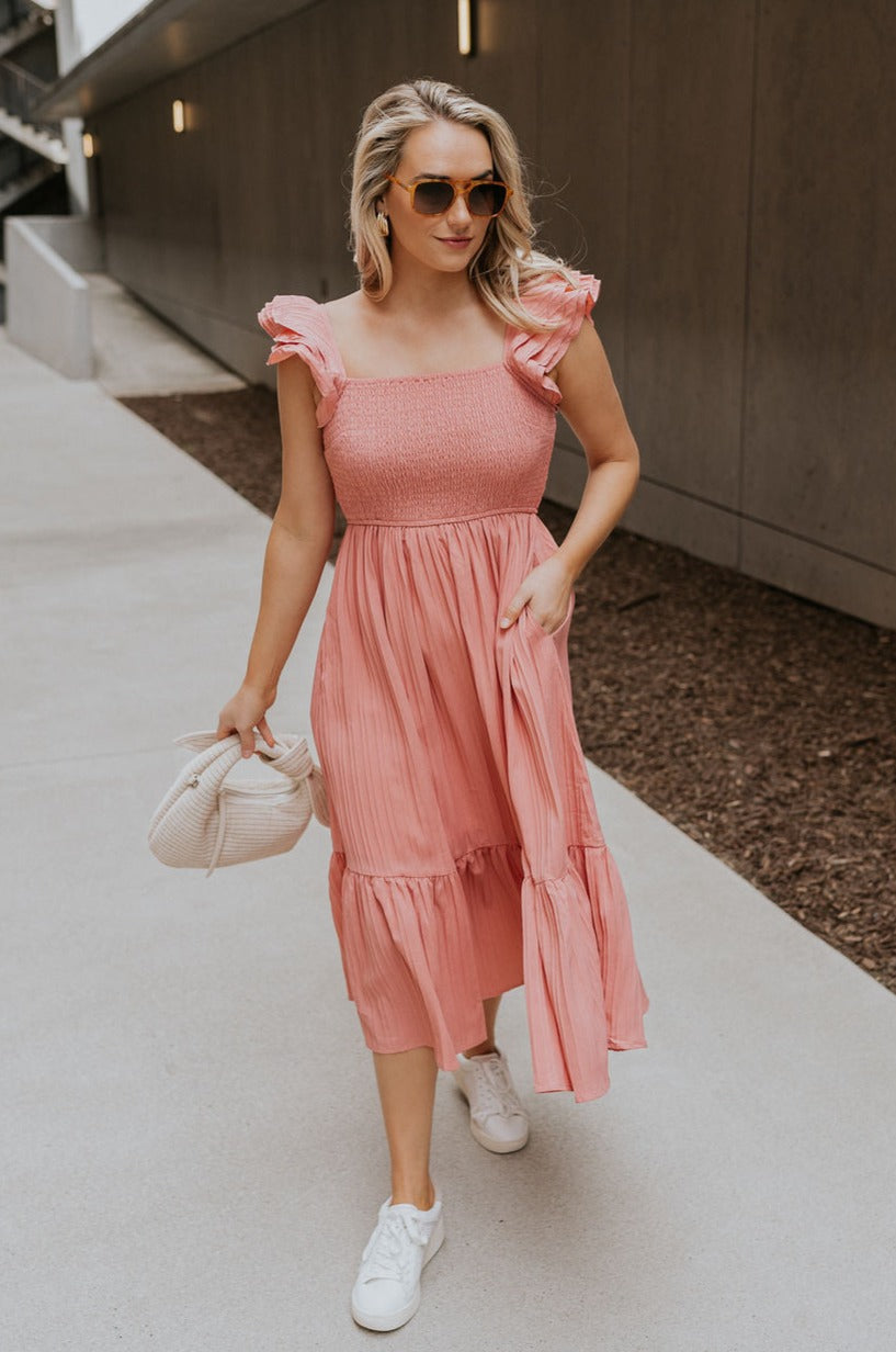 Full body view of female model wearing the Allie Coral Pink Ruffle Midi Dress which features Coral Pink Lightweight Fabric, Midi Length, Coral Pink Lining, Ruffle Tier Skirt, Smocked Upper, Square Neckline, Ruffle Straps and Two Side Pockets
