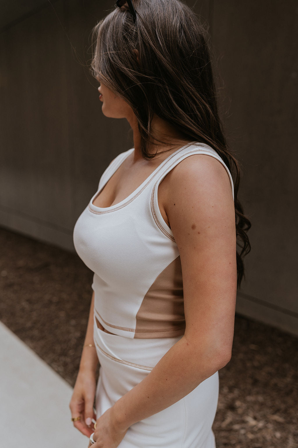 Side view of female model wearing the Hayden Cream & Mocha Tank which features Stretchy White Fabric, Taupe Side Colorblocking, Taupe Contrast Stitching, Square Neckline, Thick Straps and Cropped Waist