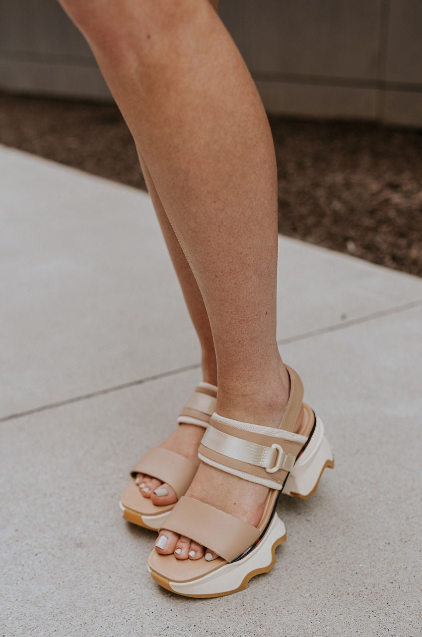 Side view of female model wearing the Kinetic Impact Slingback Heel in Honest Beige & Honest White which features Beige and Cream Leather Upper Fabric, Leather Strap, Adjustable Hook and Velcro Strap Closure, 1 1/2" Platform Sole,  2 1/12" Heel Height and Tortoise Sole Lining Details
