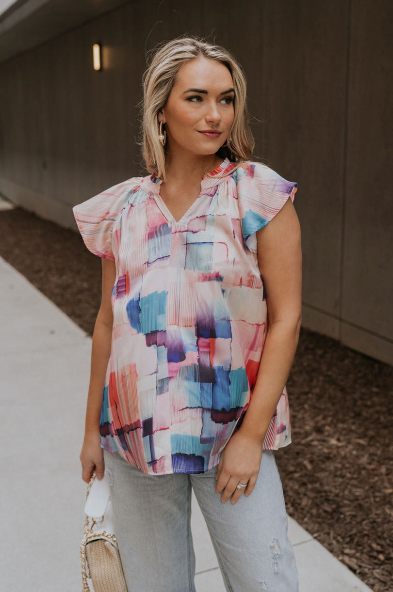 Front view of female model wearing the Vanessa Watercolor Multi Short Sleeve Top which features Satin Fabric, Purple, Blue, Peach and Pink Watercolor Block Print, V-Neckline Cut-Out and Ruffle Short Sleeves