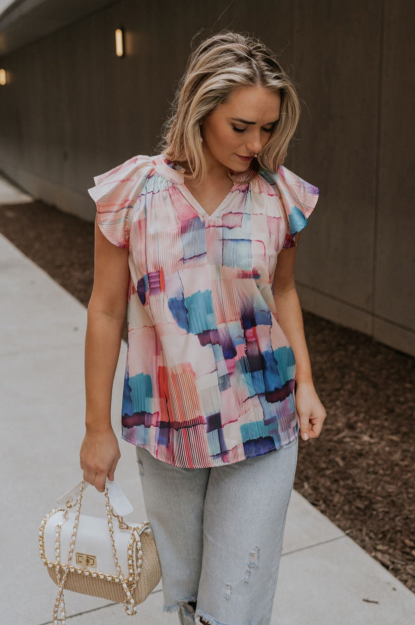 Front view of female model wearing the Vanessa Watercolor Multi Short Sleeve Top which features Satin Fabric, Purple, Blue, Peach and Pink Watercolor Block Print, V-Neckline Cut-Out and Ruffle Short Sleeves
