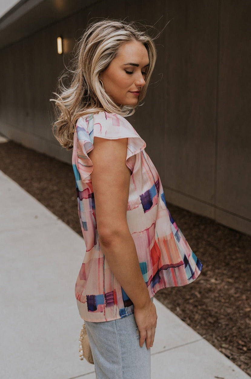 Side view of female model wearing the Vanessa Watercolor Multi Short Sleeve Top which features Satin Fabric, Purple, Blue, Peach and Pink Watercolor Block Print, V-Neckline Cut-Out and Ruffle Short Sleeves