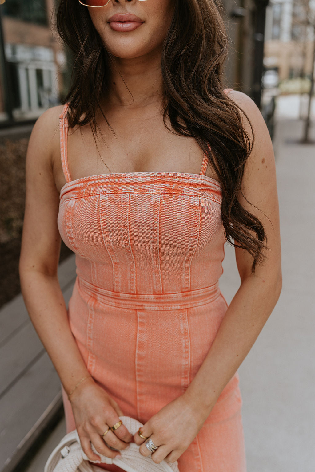 Upper body front view of brunette model wearing the Regan Wide Leg Denim Jumpsuit in Coral, that has coral denim fabric, wide legs, and thin straps. Model is holding beige purse in front of her.