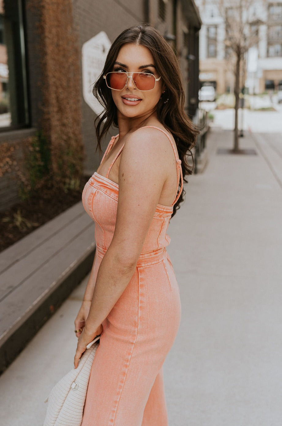 Upper body side view of brunette model wearing the Regan Wide Leg Denim Jumpsuit in Coral, that has coral denim fabric, wide legs, and thin straps. Model is holding beige purse in front of her.
