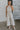 Full body view of female model wearing the Lilah Natural Linen Halter Neckline Jumpsuit which features Natural Linen Fabric, Wide Pant Legs, Tie Waistband, Twist Halter Neckline with Button Closure and  Back Key Hole Design