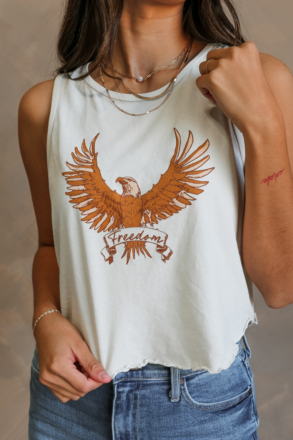 Close up view of female model wearing the Free Bird Smoke Cropped Tank which features Cream Cotton Fabric, Cropped Lettuce Hem, Round Neckline, Sleeveless and Eagle Graphic